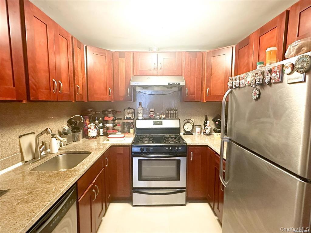 Kitchen with stainless steel appliances, light stone counters, under cabinet range hood, brown cabinets, and backsplash