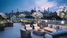 Expansive rooftop terrace with a riverfront backdrop, featuring multiple seating areas, a dining space, and integrated planters with flowering trees