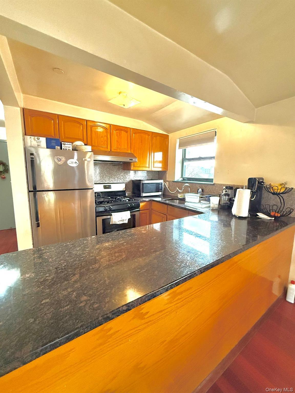 Kitchen featuring dark stone countertops, lofted ceiling, stainless steel appliances, brown cabinets, and tasteful backsplash