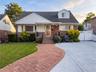 Brick and siding residence featuring a red paver walkway, mature landscaping, and a concrete driveway with a white privacy gate