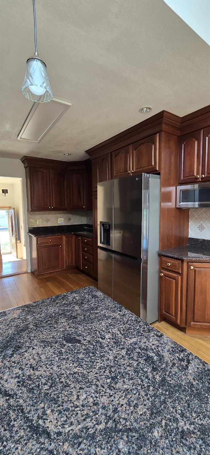 Kitchen featuring rich wood cabinetry with crown molding, stainless steel refrigerator, dark stone countertops, tile backsplash, and wood-finish floor