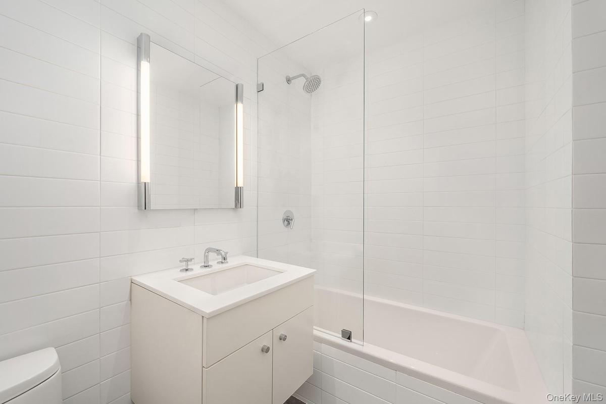 Bathroom featuring a vanity with an integrated sink and chrome fixtures, illuminated mirror with vertical lighting, floor-to-ceiling white tiled walls