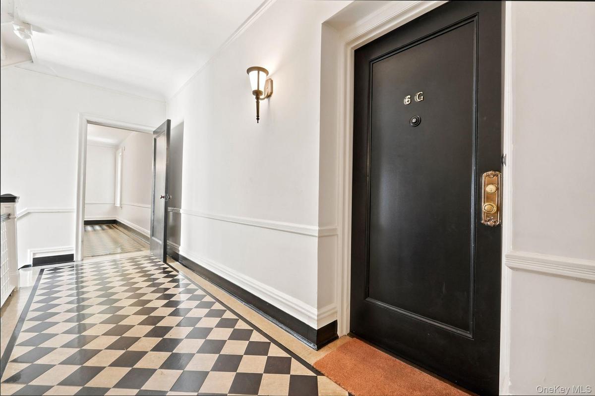 Entryway featuring checkered flooring, a black front door with brass hardware, a wall sconce, white walls with wainscoting, and a framed doorway