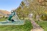 Outdoor recreational area featuring a playground structure with a green slide, a concrete pathway, a park bench, and mature trees