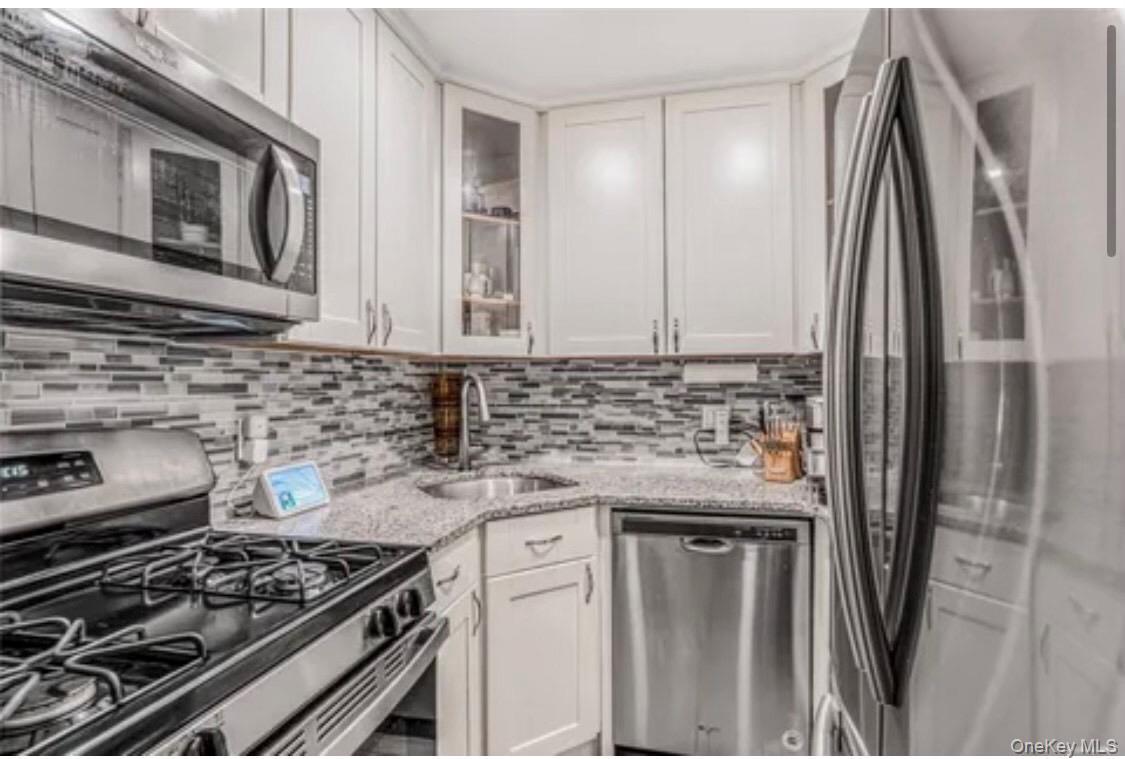 Kitchen with appliances with stainless steel finishes, glass insert cabinets, light stone countertops, white cabinets, and decorative backsplash