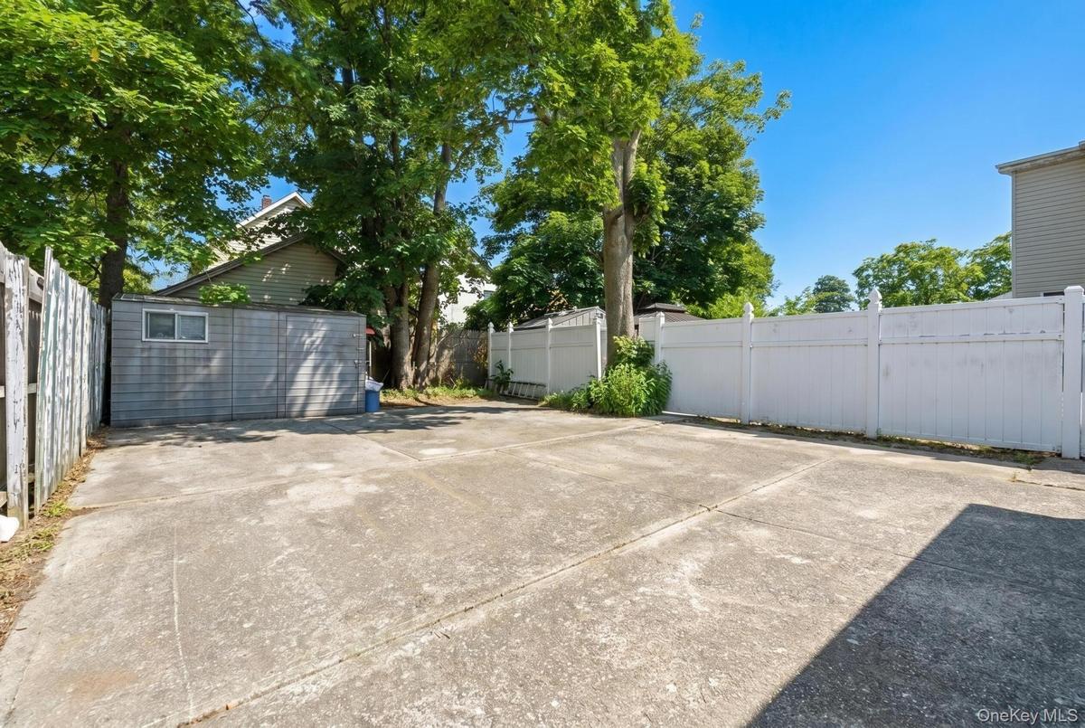 Concrete paved area featuring a detached storage shed with horizontal siding, a white privacy fence, and mature trees