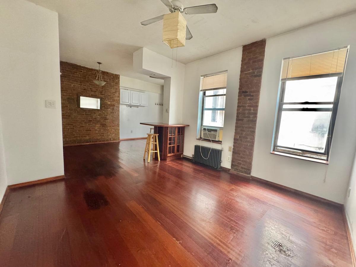 Unfurnished living room with radiator heating unit, wood finished floors, a ceiling fan, cooling unit, and brick wall