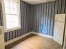 Room featuring wood-finish flooring, striped wallpaper, a double-hung window with white trim, a white baseboard, and a white interior door