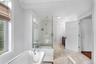 Freestanding clawfoot tub with chrome fixtures, glass-enclosed shower with marble-style tile, light-toned tile flooring, white crown molding, and a da
