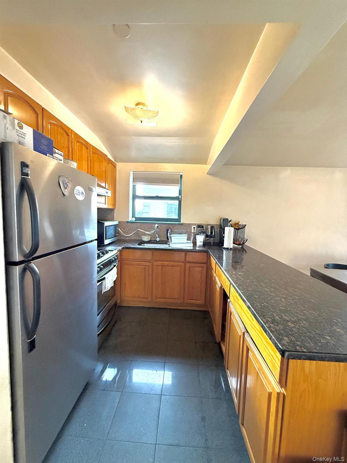 Kitchen featuring appliances with stainless steel finishes, a peninsula, dark stone countertops, brown cabinetry, and dark tile patterned floors