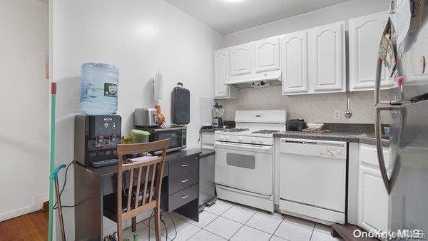 Kitchen featuring white appliances, white cabinetry, dark countertops, light tile patterned flooring, and under cabinet range hood