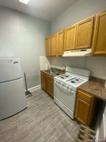 Kitchen featuring dark countertops, tasteful backsplash, a sink, under cabinet range hood, and white appliances