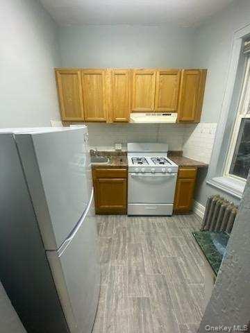 Kitchen with white appliances, brown cabinetry, baseboards, light wood-type flooring, and under cabinet range hood