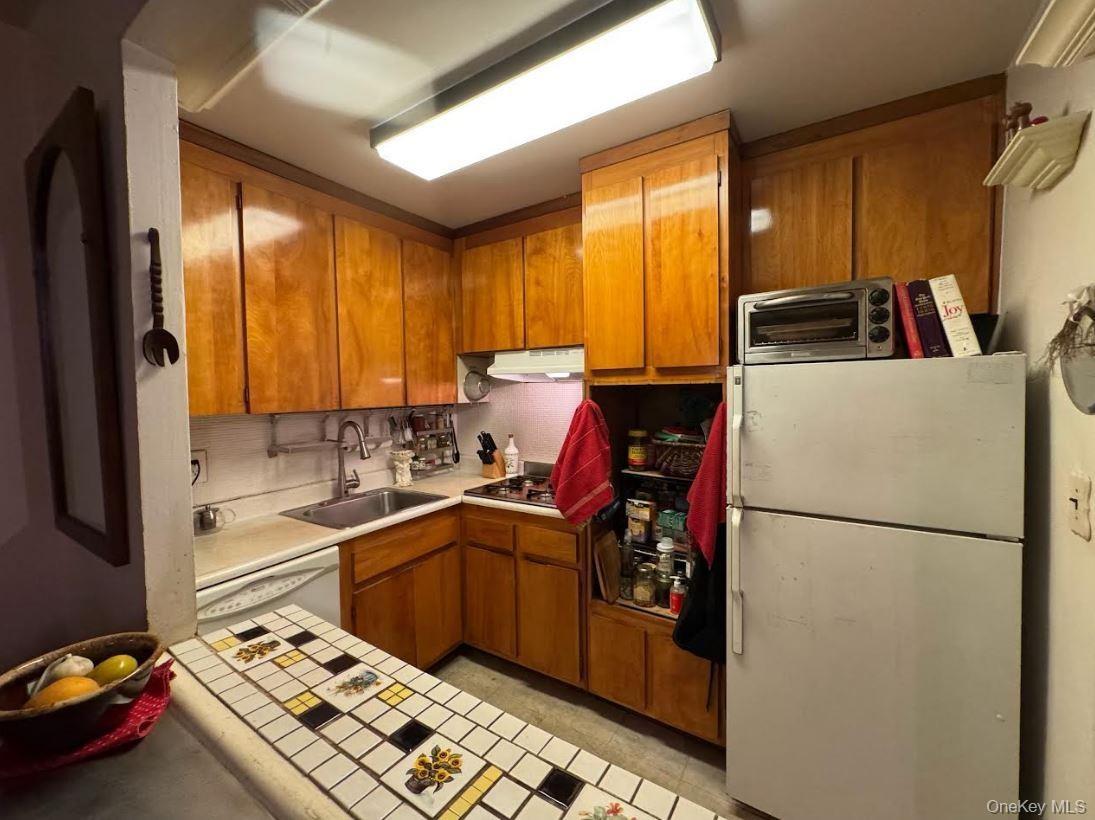 Kitchen featuring brown cabinets, white appliances, under cabinet range hood, backsplash, and tile countertops
