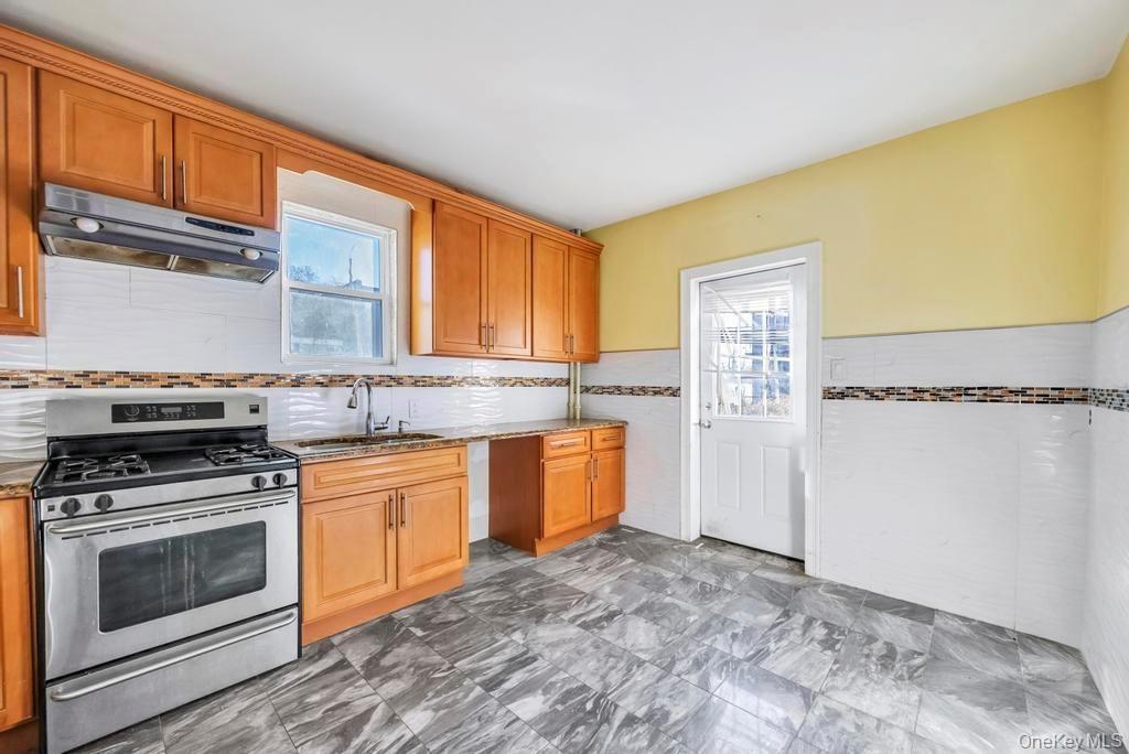Kitchen featuring stainless steel gas range, under cabinet range hood, brown cabinets, light stone countertops, and a wainscoted wall