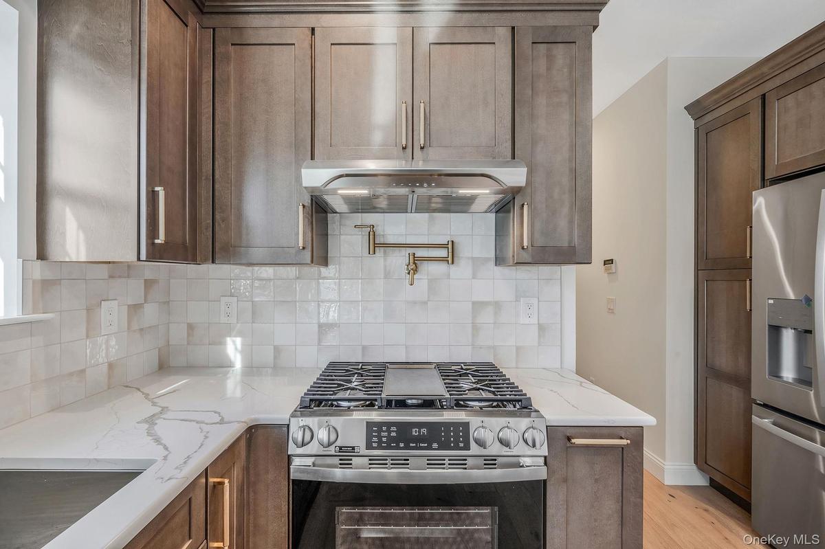 Kitchen with appliances with stainless steel finishes, light stone counters, exhaust hood, tasteful backsplash, and light wood-type flooring. For Mark