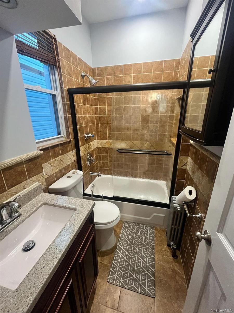 Bathroom with bath / shower combo with glass door, tile walls, vanity, radiator heating unit, and tasteful backsplash
