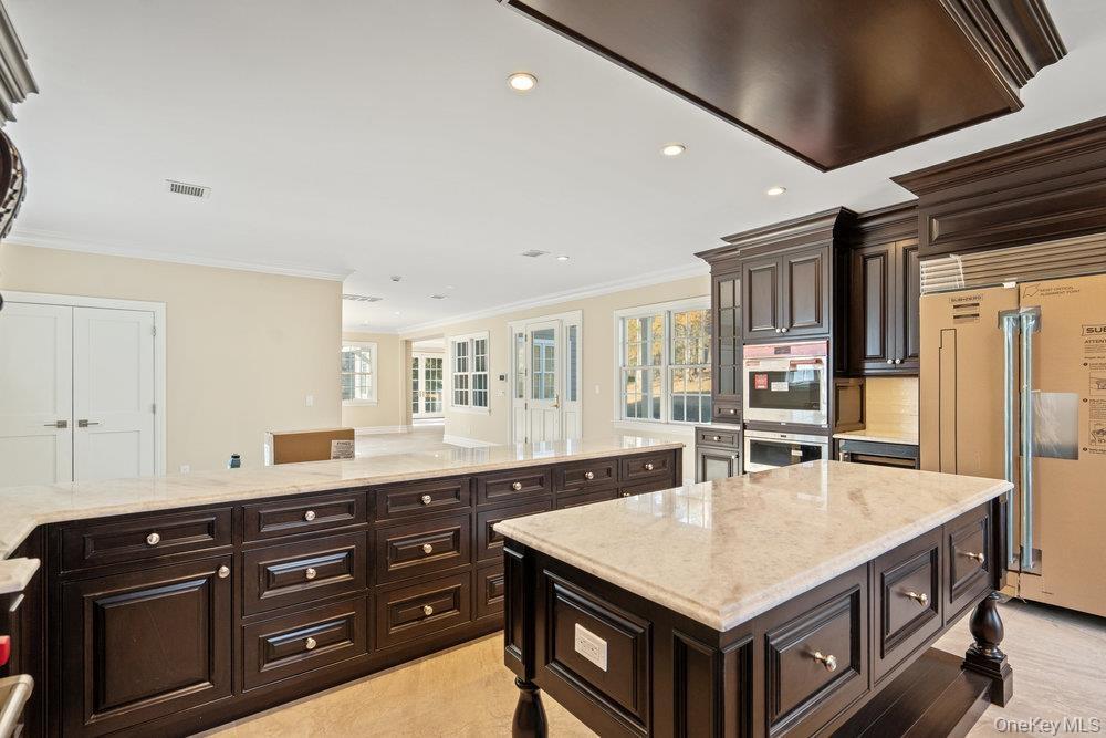 Kitchen with dark brown cabinetry, exhaust hood, paneled built in fridge, light stone countertops, and stainless steel double oven