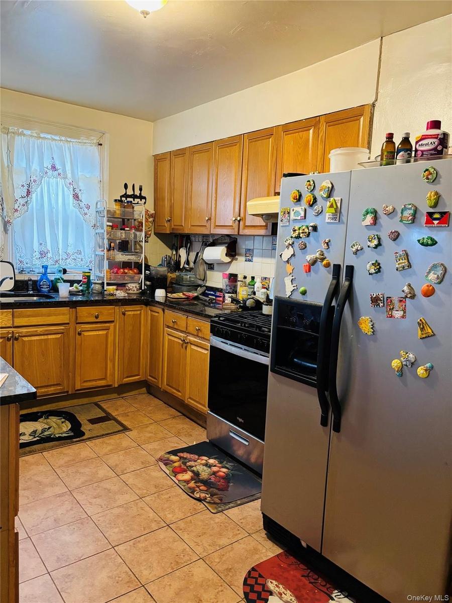 Kitchen featuring appliances with stainless steel finishes, brown cabinetry, decorative backsplash, light tile patterned flooring, and dark stone coun