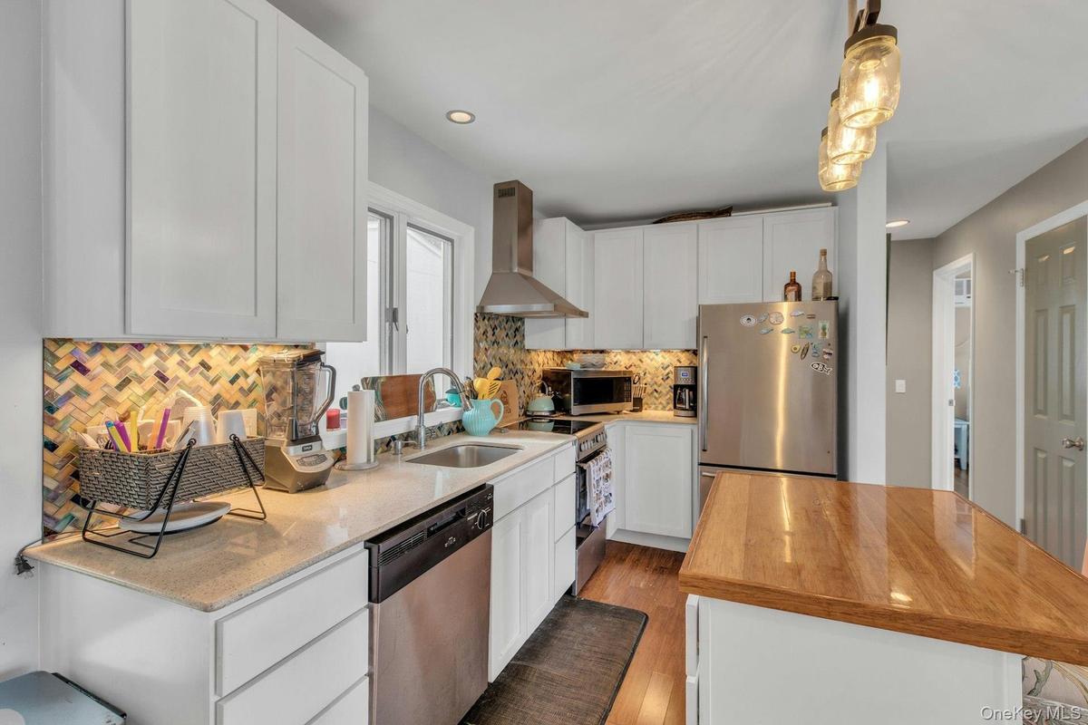 Kitchen with white cabinetry, wall chimney range hood, stainless steel appliances, light stone countertops, and hanging light fixtures