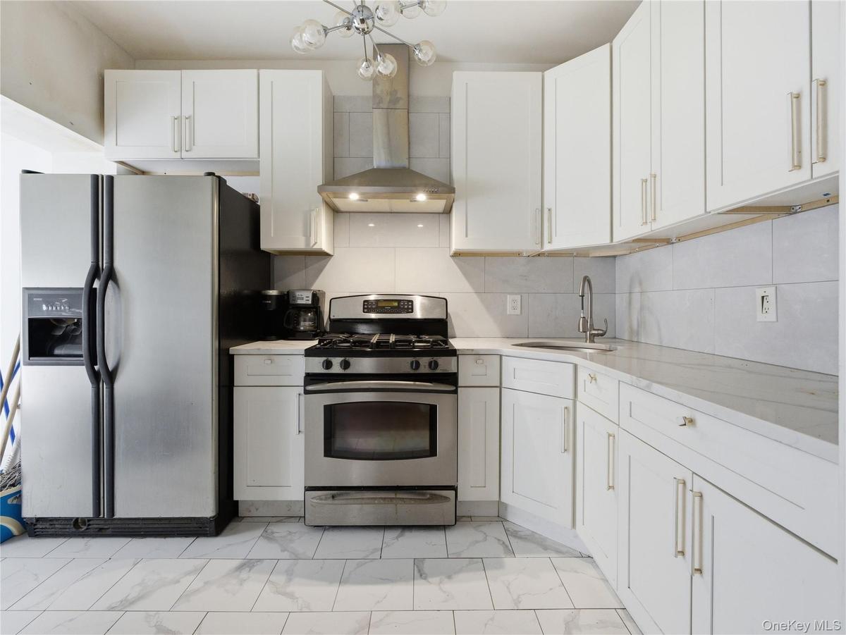 Kitchen with appliances with stainless steel finishes, light marble finish floors, wall chimney exhaust hood, and white cabinetry