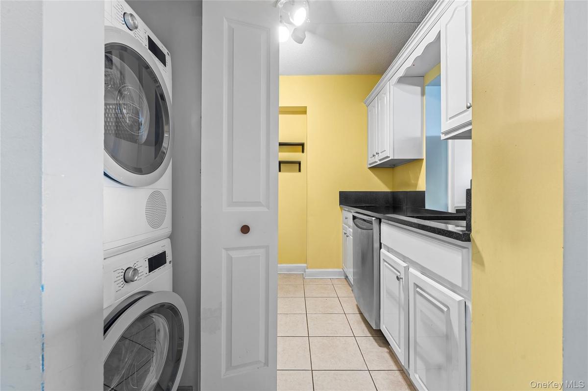Stackable washer and dryer unit, white cabinetry, black countertops, stainless steel dishwasher, and tile flooring