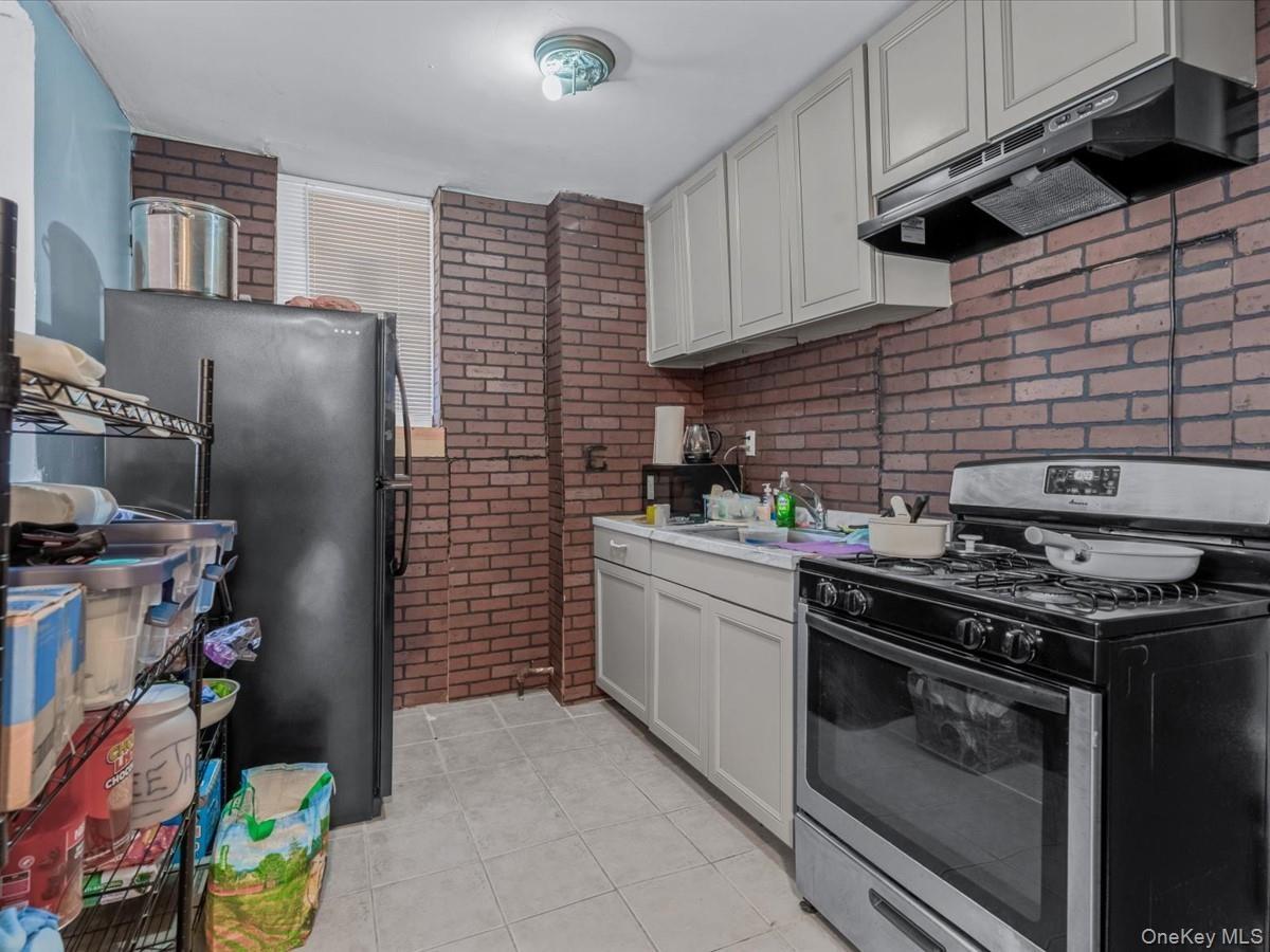 Kitchen with stainless steel range with gas cooktop, brick wall, under cabinet range hood, freestanding refrigerator, and light tile patterned floors