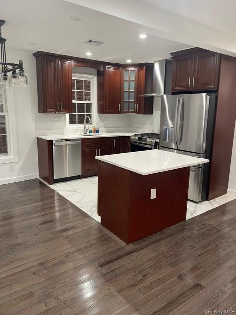 Kitchen with appliances with stainless steel finishes, light countertops, glass insert cabinets, wall chimney range hood, and a kitchen island