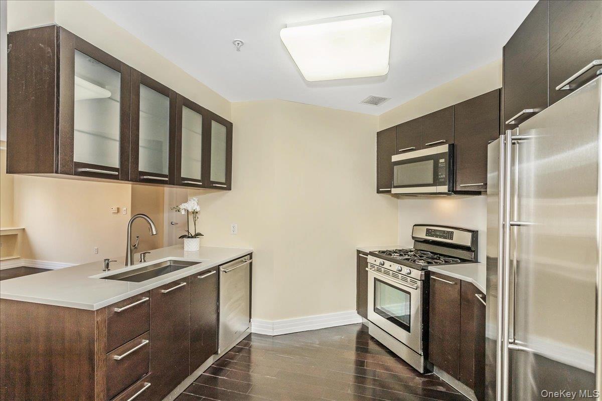 Kitchen featuring stainless steel appliances, glass insert cabinets, dark wood finished floors, and dark brown cabinets