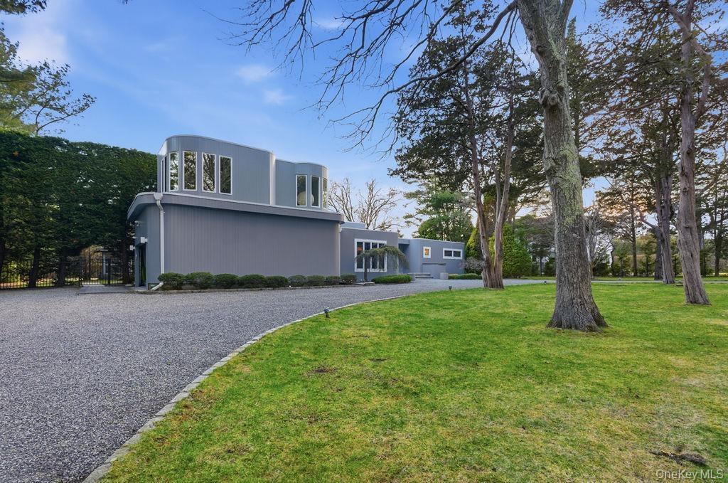 Contemporary architecture with curved walls and multiple windows, gravel driveway, and expansive lawn with mature trees