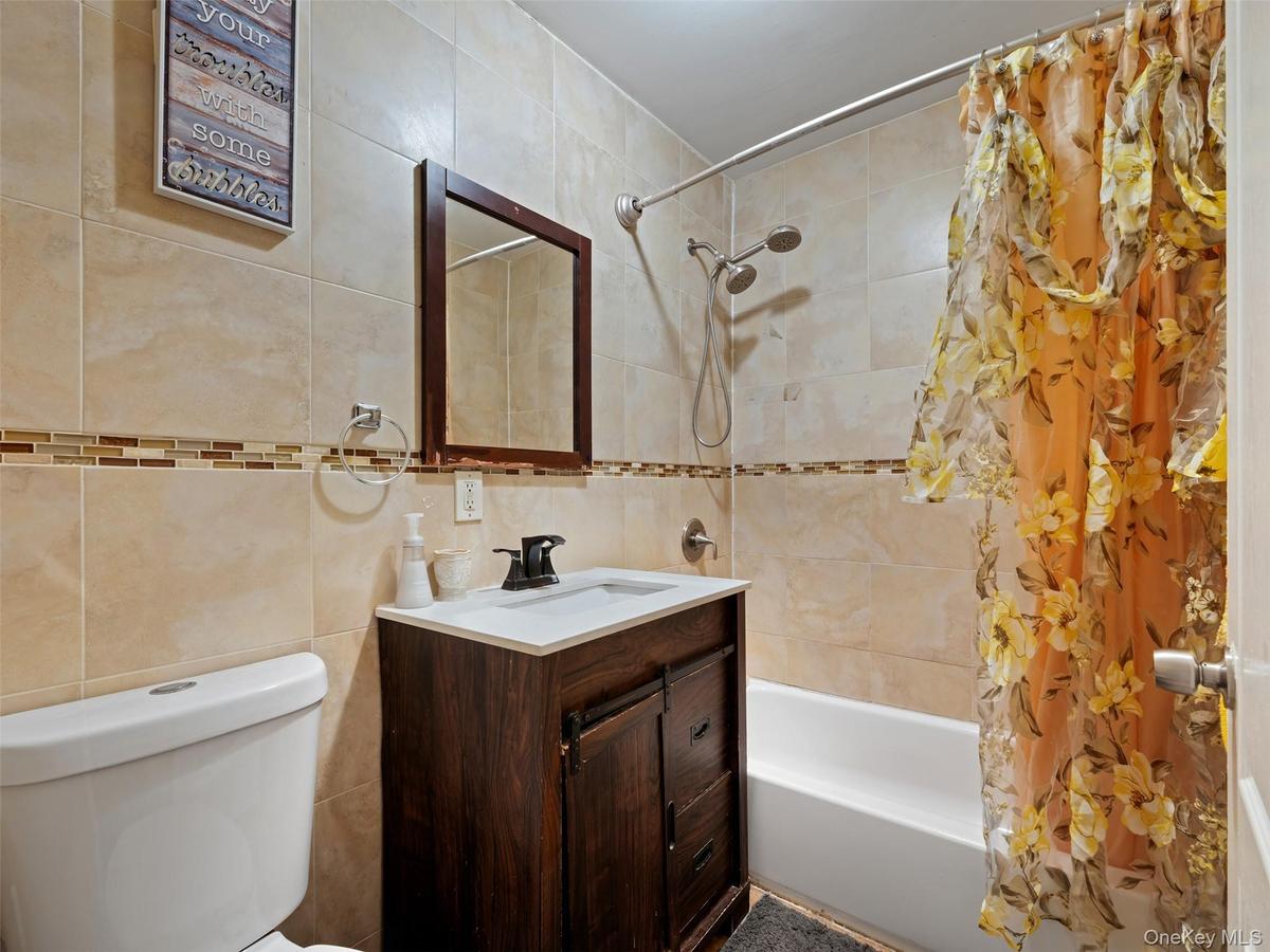 Bathroom featuring a white toilet, a dark wood vanity with a white countertop and rectangular sink, and beige tiled walls with a decorative mosaic acc