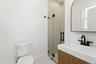 The bathroom features a modern vanity with a white countertop and dark-toned faucet, a glass-enclosed shower with tiled walls, and a white toilet