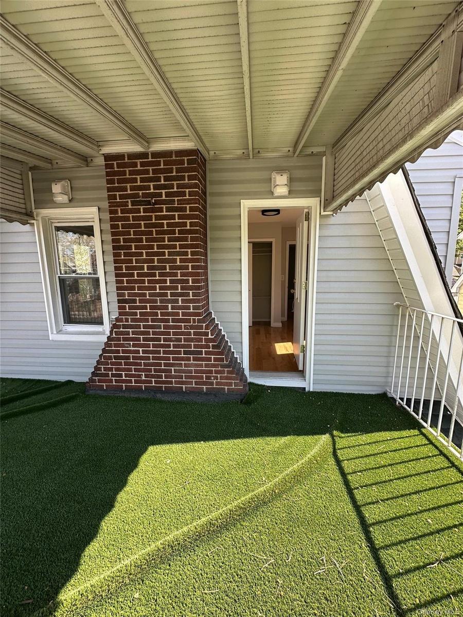 Covered porch with artificial turf flooring, featuring a red brick chimney, light gray siding, and white railing