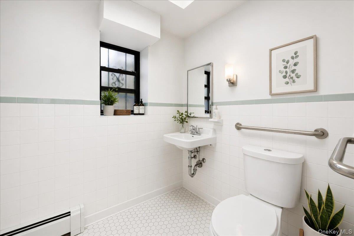 Well-lit bathroom featuring white tiled walls with a light green accent stripe, hexagonal floor tiles, a pedestal sink with exposed plumbing, a toilet