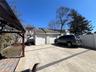 Two-car garage with white paneled doors, expansive concrete driveway with dark paver accents, and a privacy fence