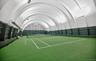 Indoor tennis court featuring a green playing surface with white line markings, an arched ceiling structure, integrated lighting fixtures, and a prote