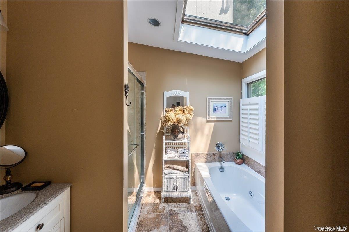 Bathroom featuring a jetted tub, glass-enclosed shower, skylight, and window with plantation shutters