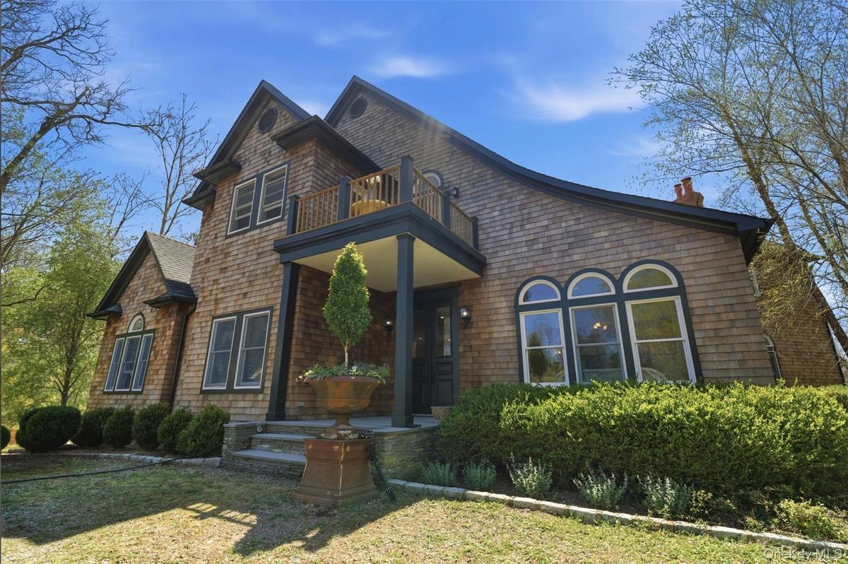 Shingle-sided exterior featuring an upper-level balcony, covered front entry with columns, multiple arched window sets, and a stone chimney