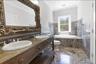 Bathroom featuring a dual vanity with vessel sinks, ornate framed mirror, integrated soaking tub, and extensive floor-to-ceiling stone tiling