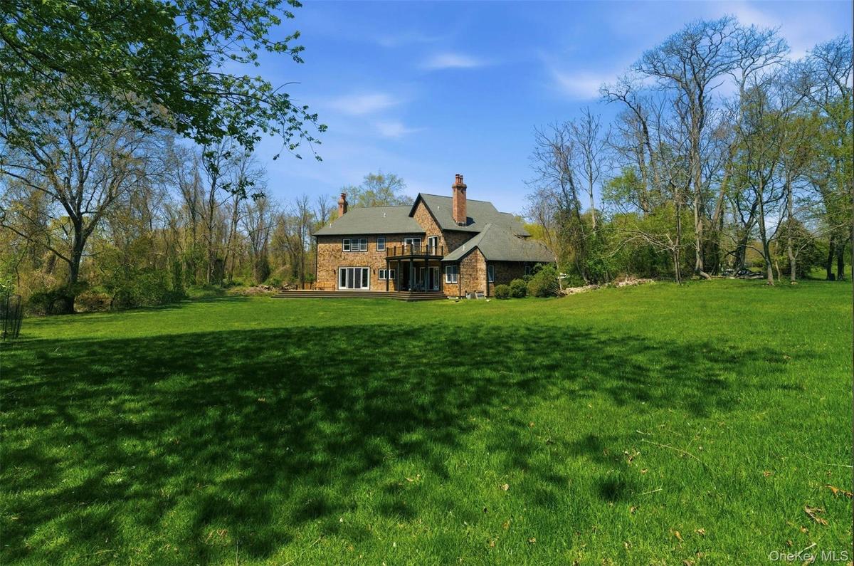 Expansive green lawn bordering a stone-clad residence with multiple chimneys, a dark shingle roof, and a rear deck featuring white-framed glass doors