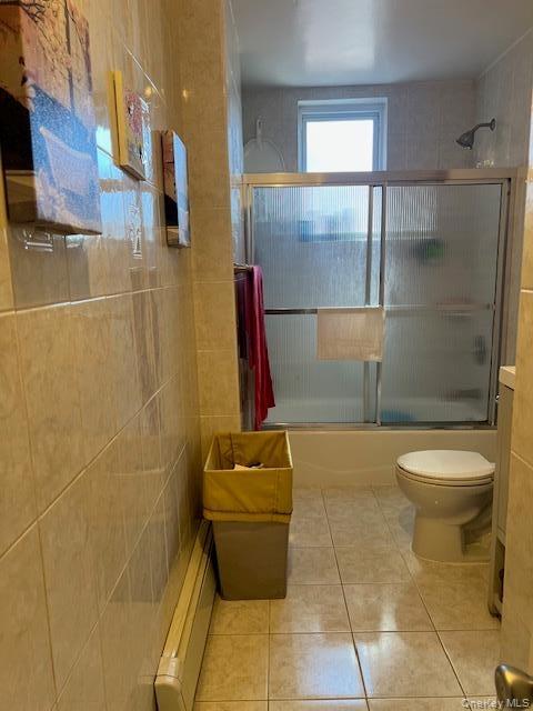 Bathroom featuring floor-to-ceiling tile, a bathtub with sliding glass doors, a window, and a standard toilet