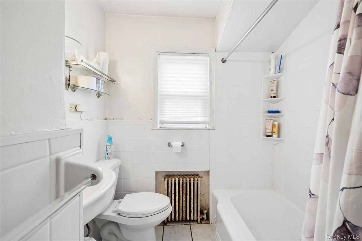 Bathroom featuring a white bathtub with a shower rod, a toilet, a window with blinds, and white wall tiling