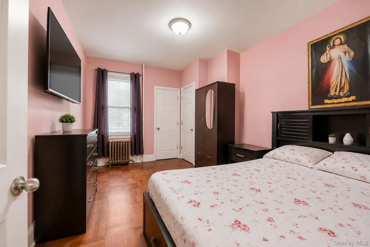 Pink-painted room featuring wood-finish flooring, a window with blinds and drapes, a wall-mounted light fixture, and a built-in radiator