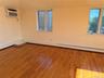 Hardwood flooring throughout featuring white baseboards, two windows with white trim, and a wall-mounted air conditioning unit