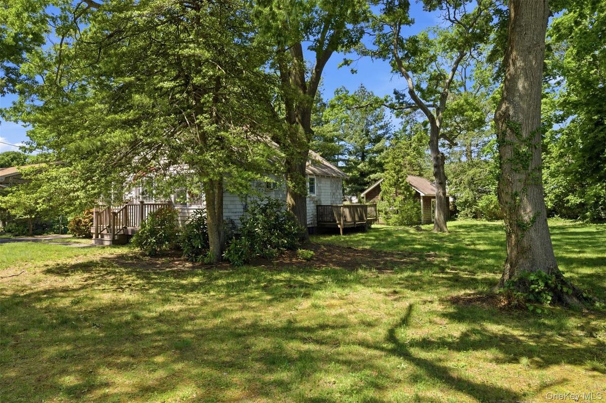 Expansive verdant lawn featuring mature shade trees, a shingled house with an exterior deck, and an additional storage shed