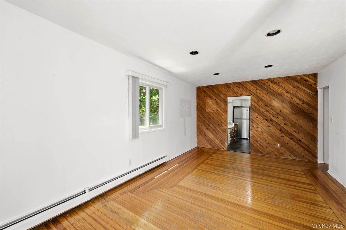 Hardwood flooring throughout, recessed ceiling lighting, baseboard heating, a single window, and a diagonal wood-paneled accent wall with an open door