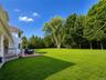 Expansive green lawn framed by mature trees, featuring a red-toned outdoor deck, white latticework, and a covered porch with white columns