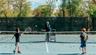 Full-size tennis court featuring a light blue playing surface, white demarcation lines, and a dark net