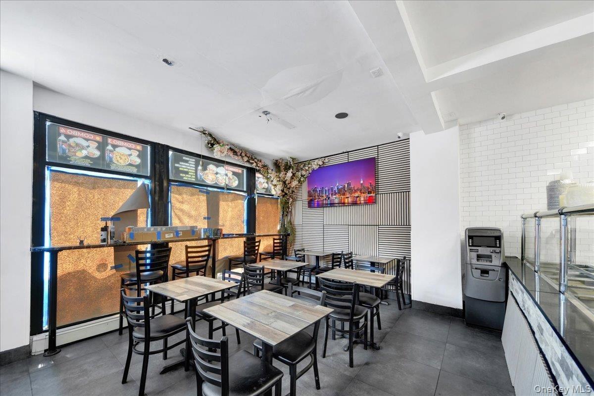 Commercial dining area featuring multiple tables and chairs, white subway tile wall, large display screen, and a service counter with glass enclosure