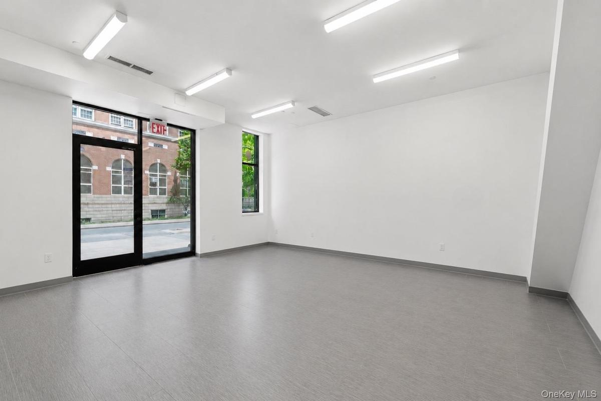 Spacious commercial interior featuring full-height storefront glass, a separate large window, recessed fluorescent lighting, light gray wood-finish fl